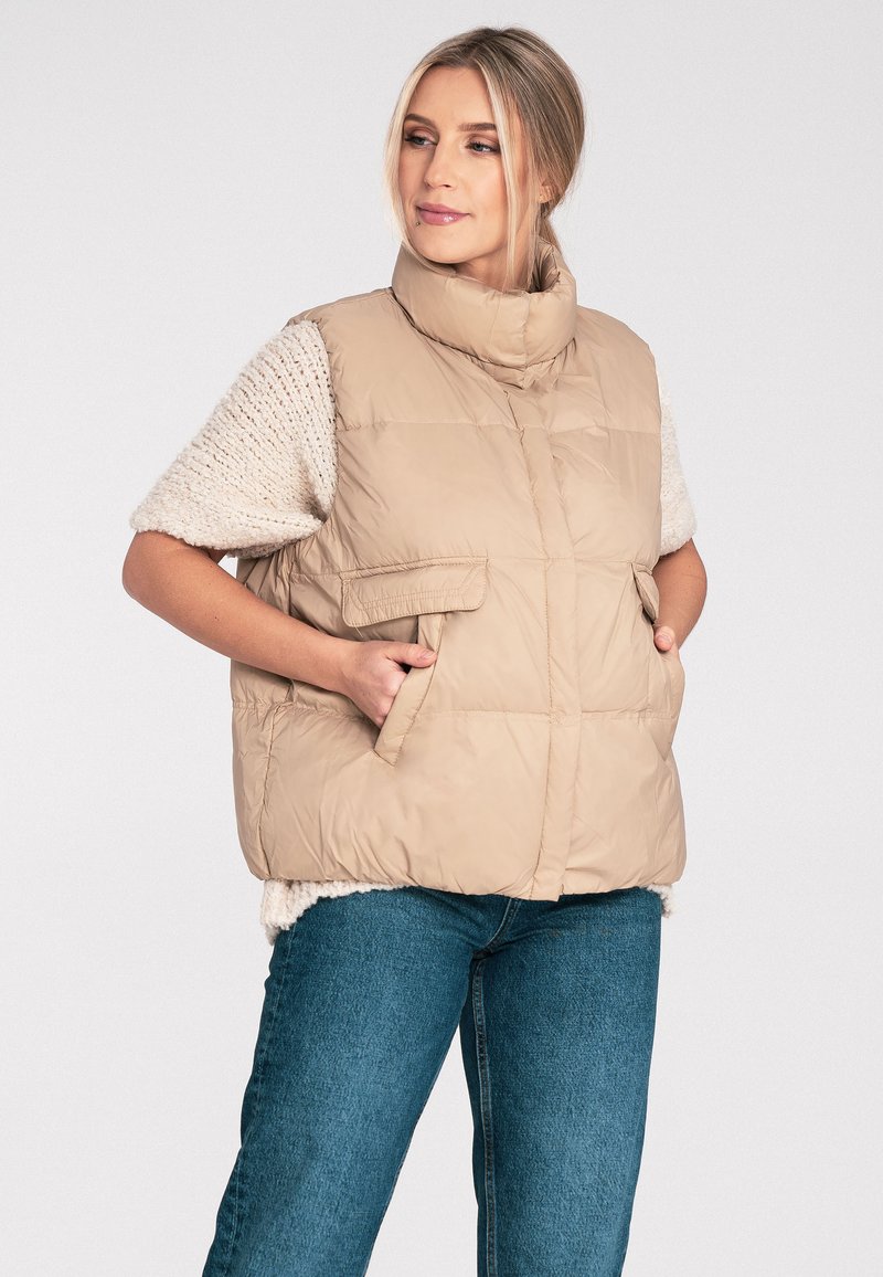 Beżowa kurtka puchowa bez rękawów z wysokim kołnierzem, dwiema przednimi kieszeniami i teksturowanymi krótkimi rękawami. Gładki, puszysty materiał z pikowanymi detalami.
