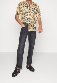 Kortærmet blomster skjorte med gule, sorte og pink mønstre, parret med mørk denim jeans og sorte vævede sandaler med tykke såler.