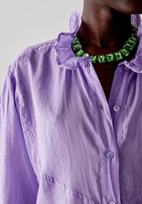 Camicia collare lilla in tessuto strutturato con chiusure a bottone, abbinata a una collana con gemma rettangolare verde.