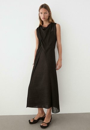 Femme debout contre un mur blanc, portant une robe maxi noire sans manches avec un col montant et des talons compensés noirs à bout ouvert.