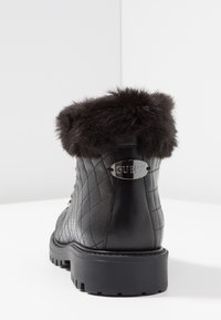 Bota negra acolchada hasta el tobillo con ribete de piel sintética, detalle de logo plateado y suela de goma. Presenta cierre de cordones en la parte frontal y parte superior texturizada.