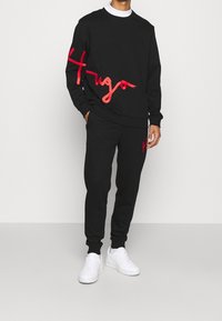 Schwarzes Sweatshirt- und Hosen-Set mit roter "Hugo"-Schrift. Weiche Baumwollmischung; Rundhalsausschnitt, gerippte Bündchen und elastischer Bund. Kombiniert mit weißen Turnschuhen.
