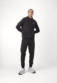 Felpa nera con tasca a marsupio e logo, abbinata a pantaloni jogger coordinati. Modello che indossa scarpe sportive bianche e nere.