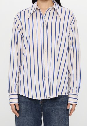 Kobieta ubrana w białą koszulę z długim rękawem z pionowymi niebieskimi paskami i niebieskie jeansy, stojąca na jednolitym tle.