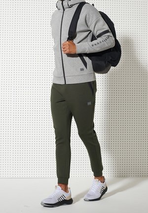 Person trägt grauen Kapuzenpullover mit Reißverschluss, olivgrüne Jogginghose, weiße Turnschuhe und trägt einen schwarzen Rucksack über einer Schulter vor einer weißen Lochwand.