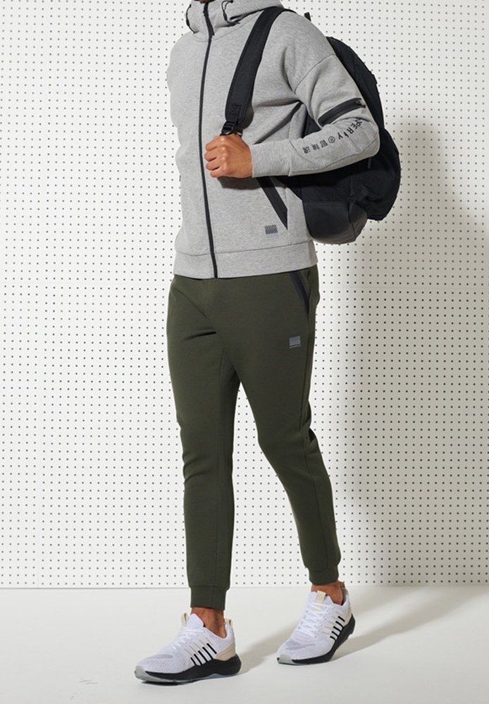 Personne portant un sweat à capuche gris zippé, un pantalon de jogging vert olive, des baskets blanches et un sac à dos noir sur une épaule devant un panneau perforé blanc.