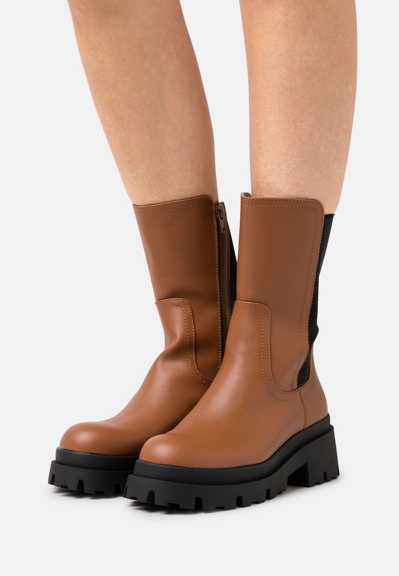 Bottes en cuir marron jusqu'au mollet avec des semelles chunky noires et une fermeture éclair sur le côté, portées sur des jambes nues devant un fond uni.