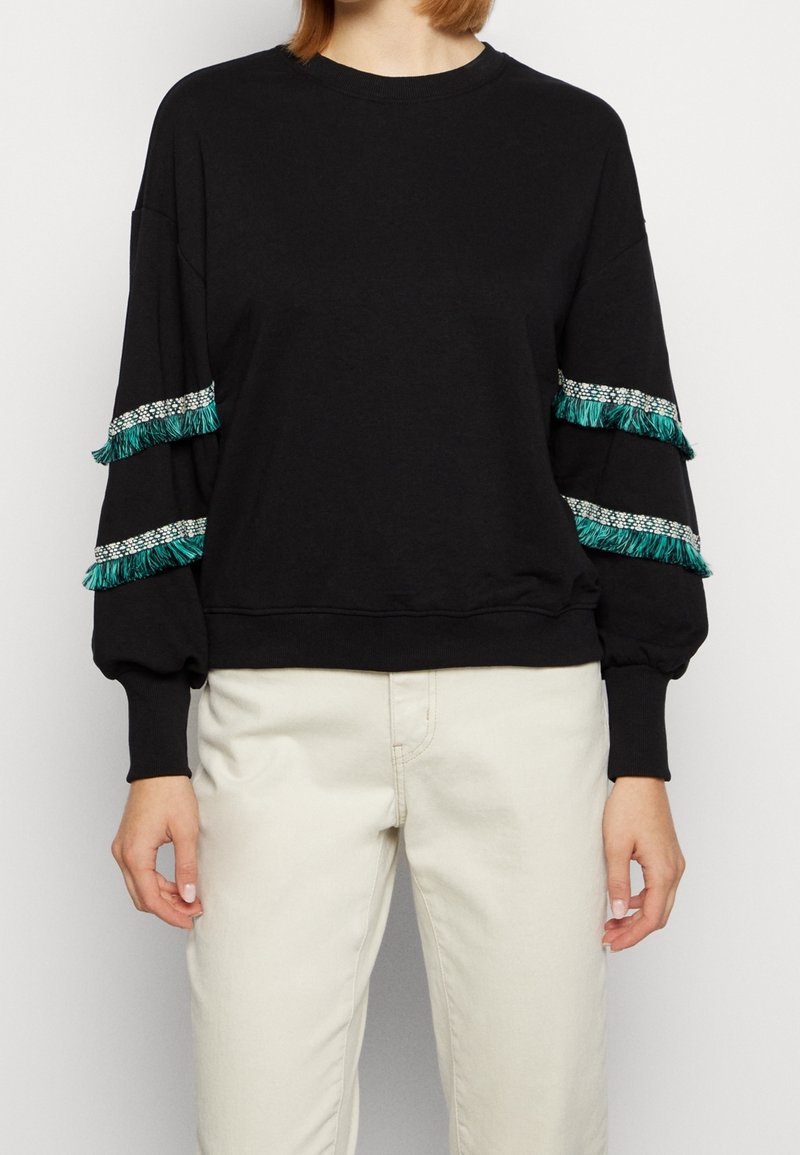 Femme portant un haut noir à manches longues avec des bandes de franges vertes et blanches sur les manches, associé à un pantalon écru.