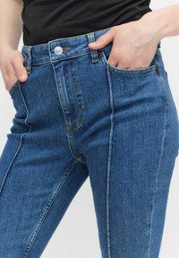 Jeans en denim bleu avec une coupe ajustée, dotés de poches avant, d'une fermeture à bouton et de coutures visibles le long des coutures.