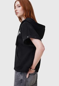 Sudadera corta negra de manga corta hecha de tela suave, presenta una gran capucha, botones de lado y un bolsillo frontal. Combinada con jeans grises holgados.