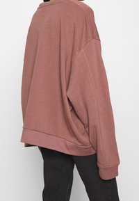 Sweat-shirt surdimensionné bordeaux avec épaules tombantes, bords côtelés et texture douce. Associé à un pantalon foncé et ajusté.