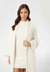 Witte, gestructureerde gebreide jurk in combinatie met een bijpassende lange cardigan, voorzien van brede mouwen en een open voorkant. Zacht, comfortabel materiaal.