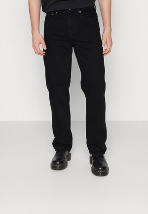 Homme portant un jean noir droit et des bottes en cuir noires, debout devant un fond clair uni.