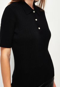 Polo noir en maille côtelée à manches courtes, doté d'un col et de trois boutons métalliques sur le devant. Texture lisse et coupe ajustée.