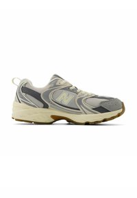 Zapatilla deportiva en gris y beige con parte superior de malla, suela de goma, lengüeta acolchada y acento del logotipo en el lateral, presentando un diseño texturizado.