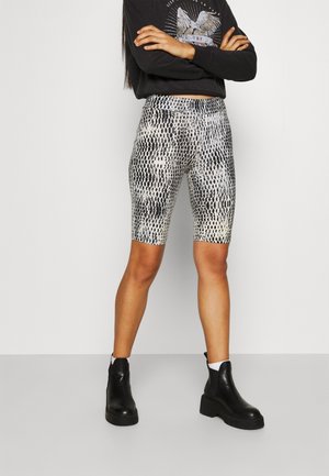 Personne portant un t-shirt noir à manches longues avec un motif graphique, un short de cycliste noir et blanc à imprimé serpent, des chaussettes blanches et des bottines noires à plateforme, debout les bras croisés.