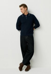 Pulover bleu marin cu fermoar, având un model texturat în grilaj, asortat cu blugi negri largi și botine din suede maro. Detaliu simplu la guler.