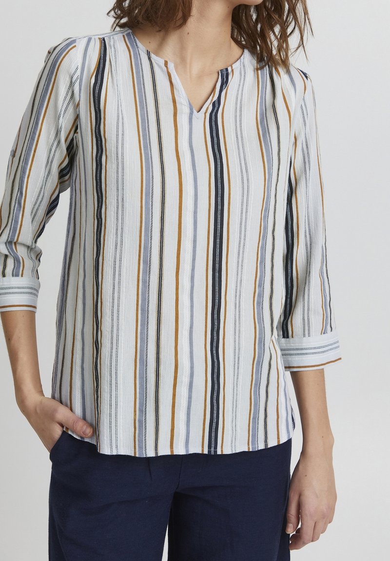 Femme portant un chemisier blanc à rayures verticales moutarde, marine et noires avec des manches trois-quarts et un pantalon marine.