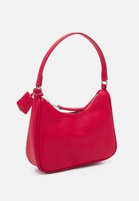Sac à main en cuir synthétique rouge avec une silhouette incurvée, une sangle courte, une fermeture éclair et une petite étiquette attachée à la poignée.