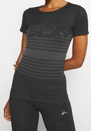 Kobieta ubrana w dopasowaną ciemnoszarą sportową koszulkę z krótkim rękawem w wzory oraz dopasowane legginsy, stojąca z jedną ręką za plecami.