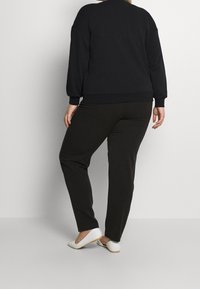 Svart sweatshirt med avslappnad passform och ribbade ärmslut, kombinerad med mörkbruna byxor och vita ballerinaskor, sedd bakifrån. Slät tyg.