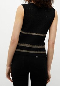 Mujer vestida con una blusa sin mangas de punto negro con rayas horizontales texturizadas y metálicas doradas, combinada con pantalones negros, vista desde atrás.