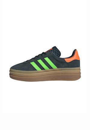 Zapatilla de ante gris oscuro con diseño de tres rayas verde neón, acento en el talón naranja y lengüeta negra texturizada, que cuenta con una gruesa suela de goma beige.