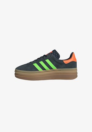 Zapatilla de ante gris oscuro con diseño de tres rayas verde neón, acento en el talón naranja y lengüeta negra texturizada, que cuenta con una gruesa suela de goma beige.