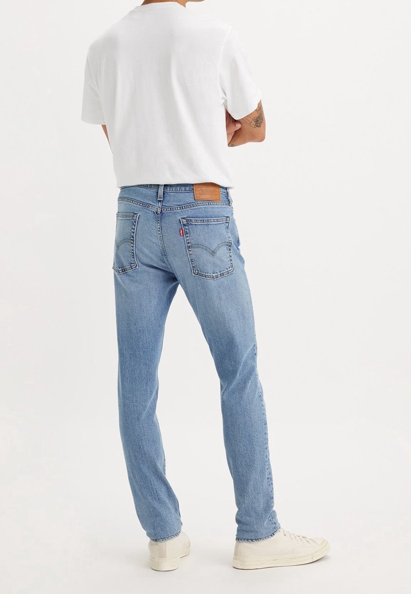 Personne portant un jean bleu clair, un t-shirt blanc et des baskets blanches, debout les bras croisés, dos à la caméra sur fond uni.