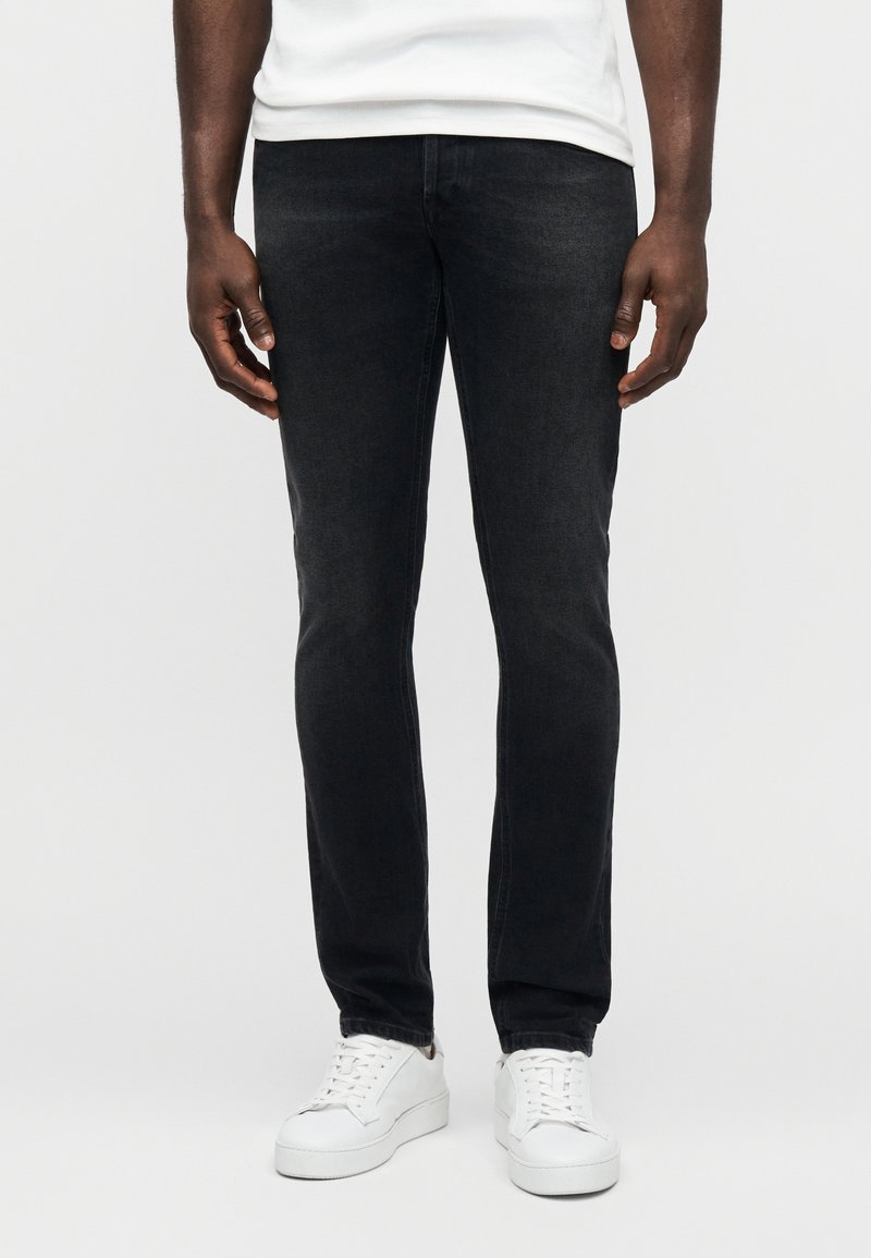 Μαύρα τζιν από τζην με στενή γραμμή, διακριτή υφή και κλασικό σχέδιο με πέντε τσέπες, συνδυασμένα με λευκά χαμηλά αθλητικά παπούτσια.