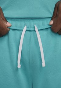 Pantalones de chándal en teal claro con cordón de ajuste con puntas blancas, textura de tela suave y cintura elástica con un detalle de costura visible.