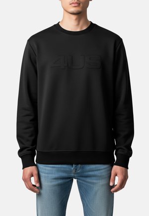 Uomo che indossa una felpa nera con logo in rilievo "4US" e jeans blu, in piedi contro uno sfondo chiaro e semplice.