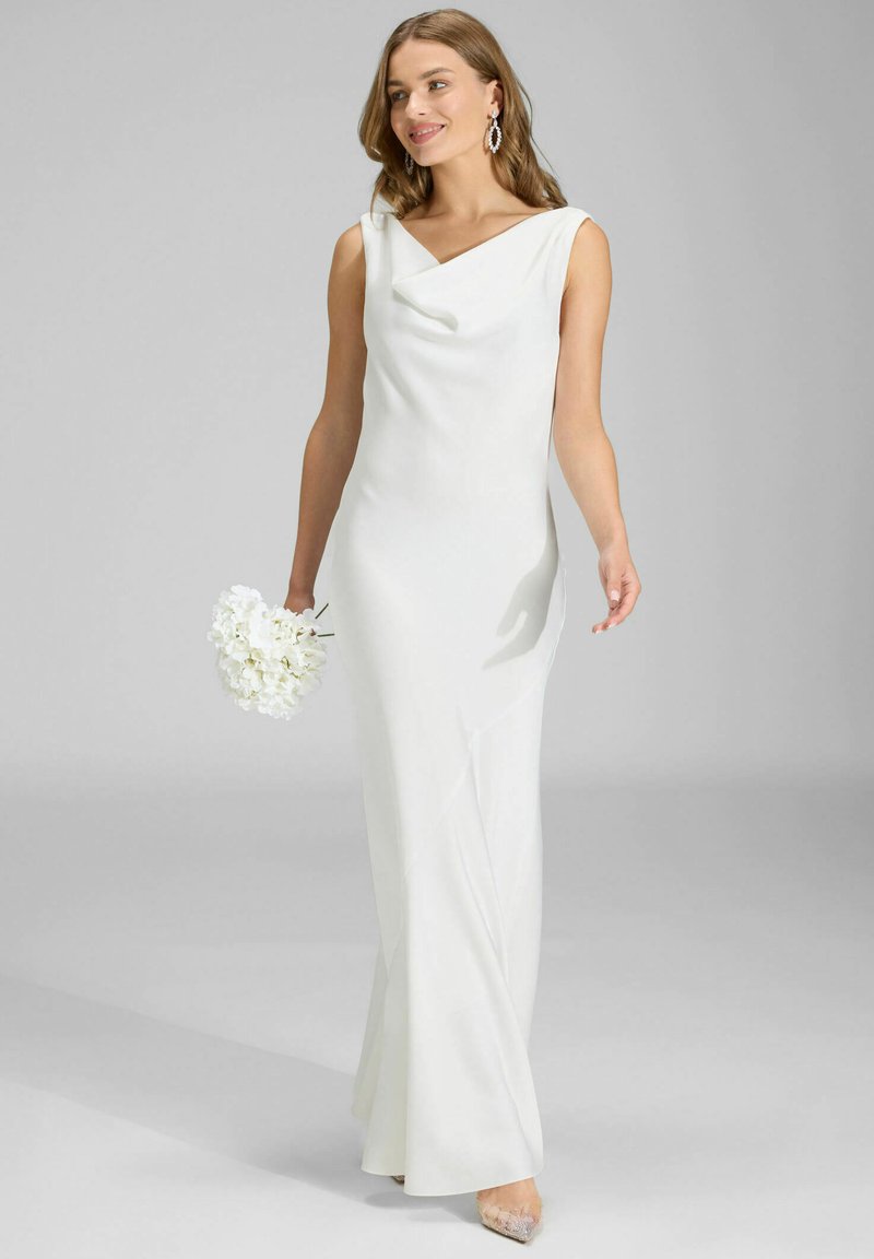 Longue robe blanche avec un décolleté drapé et un design sans manches. Tissu lisse, silhouette ajustée et fente latérale subtile ; accompagnée d'un bouquet de fleurs blanches.