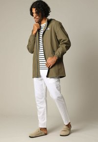 Olijfgroen lichtgewicht jack met rits, elastische manchetten, over een wit en marineblauw gestreept shirt, gecombineerd met witte broek en beige schoenen.