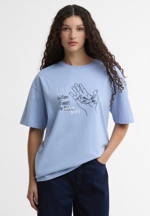 Vrouw met lang krullend haar, gekleed in een lichtblauw T-shirt met een afbeelding van een hand en een vis, met de tekst "Fortune Favours the Bold!".
