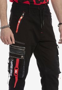 Schwarze Cargo-Hosen mit mehreren Reißverschlusstaschen und Nieten. Merkmale sind rote Akzente und ein elastischer Bund mit Kordelzug. Das Material wirkt langlebig.