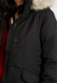 Parka noire avec capuche doublée de fausse fourrure, dotée d'une fermeture éclair et de détails à boutons-pression, et d'une poche latérale. Couche intérieure rouge visible.