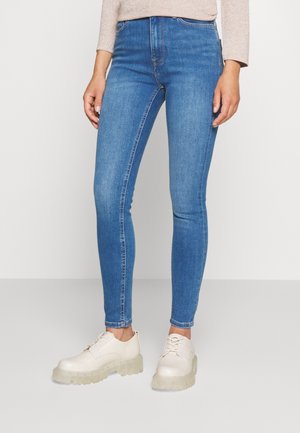 Personne portant un jean skinny bleu clair et des chaussures à lacets épaisses blanc cassé, debout devant un fond uni.