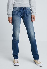 Donkerblauwe denim jeans met een rechte pijp, lichte vervaging en minimale slijtage. Gecombineerd met lichtbruine sneakers en een grijze sweater.