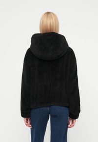 Μαύρο μπουφάν με κουκούλα, κατασκευασμένο από απαλό, υφή υφασμένο ύφασμα. Διαθέτει μακριά μανίκια, άνετη εφαρμογή και τελείωμα στη μέση.