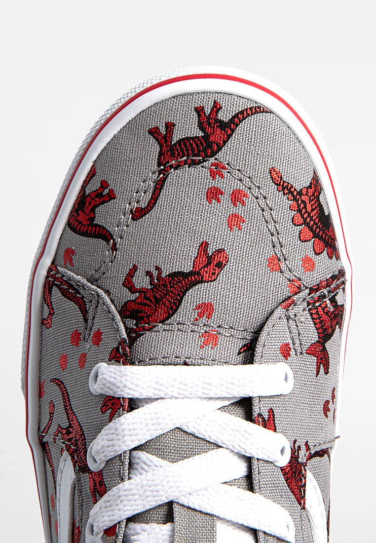 Zapatilla de lona gris con estampado de dinosaurios rojos y huellas de patas. Cierres blancos y suela de goma con una banda de acento roja.