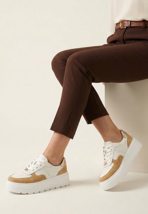 Persoon zit, draagt een bruine broek en witte sneakers met lichtbruine suède details en dikke witte zolen.