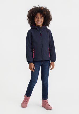 Mädchen mit lockigem Haar, das eine marineblaue Jacke mit pinken Reißverschlüssen, dunkle Jeans und pinke High-Top-Sneaker trägt, lächelt und vor weißem Hintergrund steht.