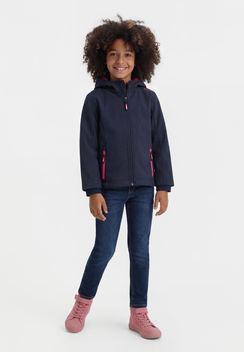 Κορίτσι με σγουρά μαλλιά, φοράει μπλε σκούρο μπουφάν με ροζ φερμουάρ, σκούρο τζιν και ροζ αθλητικά παπούτσια υψηλού πάνω, χαμογελώντας και στέκεται μπροστά σε λευκό φόντο.