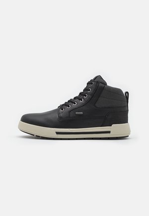 Zapatilla de deporte negra de piel alta con suela de goma beige, cordones delanteros, cuello acolchado para el tobillo y lengüeta en fondo blanco.