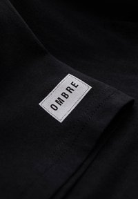 Tissu noir avec une étiquette rectangulaire blanche cousue près du bord, affichant le nom de la marque « OMBRÉ » en lettres majuscules noires.