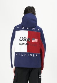 Farveblokeret vindjakke i marineblå, rød og hvid. Har "USA SAILGP TEAM" logo og "TOMMY HILFIGER" tekst i kontrastfarver. Hætte-design.