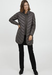 Tamsiai pilkas pūstas ilgas paltas su siuvinėtu raštu, aukšta apykakle ir centru užtrauktuku. Dėvimas virš prigludusio viršūnės ir juodų kelnių.