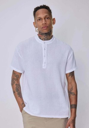 Homme avec des tatouages sur le cou et les bras, portant une chemise blanche à manches courtes boutonnée et un pantalon beige, debout, une main dans la poche.