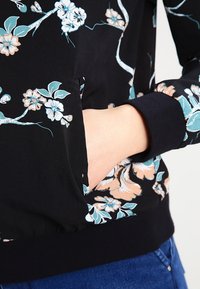 Tissu floral noir avec motifs aqua et pêche. Comprend une poche latérale, des poignets côtelés et un ourlet. Mettant l'accent sur la texture tactile et les détails du design.